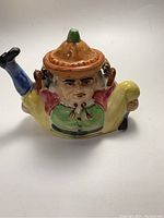 Side view of multi-colored Japan ceramic Toby style teapot, showing figure in traditional clothing, blue shoe spout, body with green and pink colors, and yellow pants.