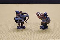 Two figurines shown from the front on a beige background, the archer with bow and the warrior with mace and shield visible.