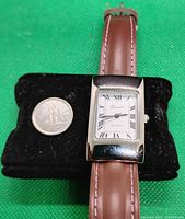 Front view of unisex quartz wristwatch with brown leather strap next to a quarter for scale. Silver-tone rectangular case and white dial with Roman numerals visible.