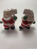 Front view of two Santa Claus salt and pepper shakers showing facial details and holiday signs held in hands.