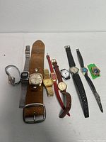 Full view of all ten vintage Timex watches laid out on white surface varying in strap material and dial face shapes and colors