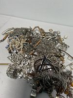 Pile of mixed jewelry showing various silver-tone chains, pendants, and shell/pearl beaded necklaces laid on white surface