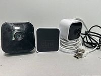 Front view of two Blink home security cameras and a Blink chime box with cords on a white background.