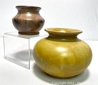 Two vintage pottery vases displayed, one yellow glazed Blue Mountain pottery and one brown glazed Huronia pottery, shown on an acrylic stand.