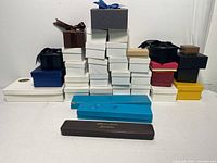 Front view of assorted cardboard jewelry boxes stacked together, showing variety in size, shape, and color including white, black, blue, yellow, brown, and red/pink boxes, some with ribbons.