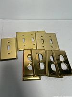 Eight solid brass electrical outlet and switch covers arranged on a white surface. Includes four switch plates and four outlet covers.