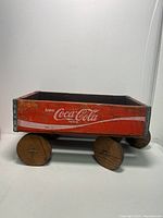 Side view of the red wooden cart with Coca-Cola branding and visible wear, showing metal corner reinforcements and round wooden wheels.