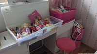Clear plastic storage bin on table containing multiple Barbie and other fashion dolls, decorative white metal doll chair with pink cushion next to it, pink storage tub filled with accessories behind.