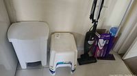 Photo showing white plastic pedal garbage bin, two step stools (one plain white, one white with blue accents), and the upright vacuum cleaner and Swiffer Wet Jet boxed unit against wall.
