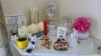 Overview of assorted decorative items on a corner shelf including candles, figurines, a white cat figurine in yellow pot, small glass vases, and a white vase with pink artificial flower.