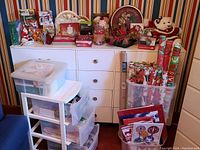 Wide view of white plastic drawer unit with assorted Christmas decor, gift wrap, and cards on top and in clear storage bins beside it.