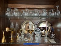 Lower shelf showing crystal decanter with stopper, two engraved silver plate trays, silver plate bells with decorative tops and additional glassware.
