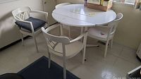 White round dining table with four white chairs around it, one chair with dark blue cushion, the table shows signs of scratches and has two leaves.