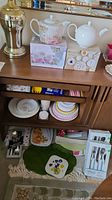 General overview of assorted dishes and some flatware with boxed items on shelf and drawer below.