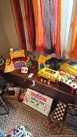 Photo showing a collection of vintage toys including a Fisher-Price Grand Piano, Monopoly game board, toy bus, and assorted smaller toys on a table near window.