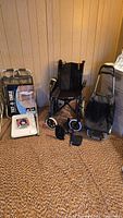 Full view of the wheelchair, walker, grocery trolley, and tilt table with accessories placed on the floor against wood panel walls.