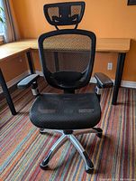 Front view of black mesh Staples Hyken ergonomic office chair with headrest, armrests, and mesh seat, photographed in room with orange walls and striped carpet.