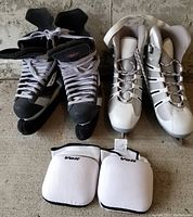 Black and gray men's ice skates size 8, white women's ice skates size 10, and white knee pads on a rough textured floor.
