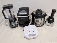 Front view of all assembled appliances: blender with pitcher, ice maker, pressure cooker, frozen treat maker, sandwich maker.