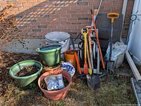 Overview of landscaping supplies including planters with dirt, garden tools, gravel and stone dust bags, garden gloves, and garden hose against brick wall.