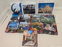 Photo showing eleven boxed jigsaw puzzles on floor, featuring various scenes and counts of pieces (mostly 1000 pieces)