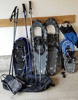 Photo showing 3 pairs of snowshoes, 4 walking poles, 2 gaiters, and a snowshoe bag arranged against a wall.