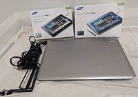 Photo showing two retail boxes for Samsung Galaxy Tab 2 10.1 and Galaxy Note 10.1, a silver HP Pavilion laptop closed, power cables and tablet stands lying on a white surface