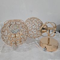 Gold modern wall light with spherical cage having crystal-like circular elements and LED light inside, gold base and support arm.