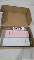 Two mini heat sealers, pink and white, packaged in a cardboard box with a white USB charging cable coiled above them.
