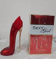 Photo shows full view of the Sexy Girl Eau de Parfum bottle shaped like a red high heel shoe with metallic gold heel next to its original boxed packaging which is red and white with gold trim.