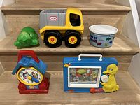 Photo showing Fisher Price wind-up musical grandfather clock, Sesame Street music box with Big Bird and TV screen, Little Tikes yellow dump truck, and a plastic tub filled with small toy animals.