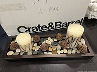 Photo shows a Crate & Barrel branded rectangular wooden tray containing eight white pillar candles with dark marbled patterns. The base is decorated with natural stones and pinecones arranged evenly throughout.