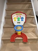 Photo showing Fisher Price children's basketball hoop stand with attached colored targets and small hoop, placed on wooden stairs.