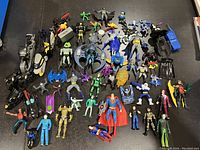Full view of large collection of DC action figures and vehicles arranged on a black surface including Batman, Superman, Joker figures and several Batmobile models.