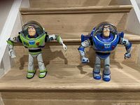 Two Buzz Lightyear talking toys, one green and one blue, with arms posed on wooden steps
