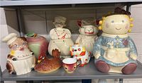 Shelf with eight vintage ceramic cookie jars and related pieces in various animal and character shapes including pig chef, large doll, apple jar, hen, cat/lion, and small cup.