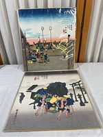 Photo showing both vintage Japanese woodblock prints, one wrapped in plastic and standing upright, the other laid flat on the table, depicting scenes from old civilizations.