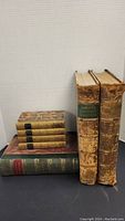 Front view of seven antique books showing spines and covers. Four smaller volumes with marbled covers likely John Dryden works. Two large worn leather-bound volumes of House of Commons Debates and one large volume labeled Cassels Family Magazine 1881.