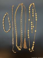 Five gold-colored costume jewelry pieces: four long necklaces and one bracelet, arranged on black background.