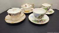 Four vintage cups and saucers displayed on a black surface in a box with white backdrop, showing varying floral and gold designs