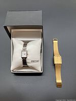 Two Seiko watches shown, one ladies two-tone watch in box with pillow, one men's gold-tone watch next to it, grey background.
