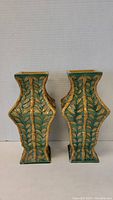 Front view of two green vases with raised bamboo leaf motifs in yellow, square base, 10 inches tall each.