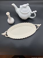 White teapot with sculpted bird lid on a black background, ivory oval platter with bird handles, and slender white flower vase.