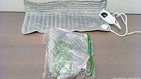 Two plush electric heating mats with gray soft fabric surface. One mat is displayed flat with attached wired controller showing heat and timer settings. The other is packaged in clear plastic bag.