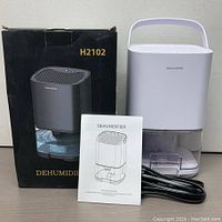 Photo showing the white dehumidifier with handle, transparent water tank, black power cord, instruction manual, and original black product box labeled H2102.