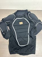 Black Benken outdoor body armour jacket laid flat showing padded protective armor panels on chest, back, shoulders, and sleeves with silver reflective piping.