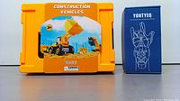 Orange plastic case labeled 'Construction Vehicles' with a picture of a dump truck and construction background and a blue box labeled 'Yontyeq' with instructions on hand use.