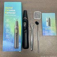 All items included in the lot laid out on a wooden surface: the black handheld dental calculus remover, multiple metal tool heads in a small plastic box, dental mirror, metal hook, dental scraper, user manual, and original packaging box.
