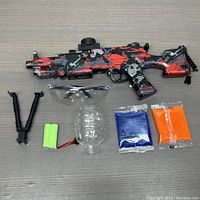 Gel ball blaster toy in red and black camo with accessories including bipod, battery, water reservoir, safety glasses, and two gel ball packets (blue and orange).