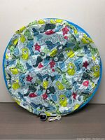 Top view of round interactive cat toy cover with colorful monster patterns and blue edges, including a small motorized toy with a white USB charging cable placed near it.